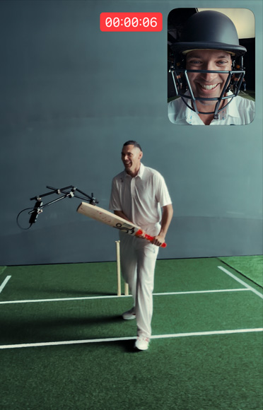 Cricketer Scott Boland stands in front of a wicket on an indoor cricket pitch holding a bat with iPhone 17 Pro attached, ready to strike. In the top right-hand corner, a picture-in-picture window shows a selfie view of cricketer Alex Carey wearing a helmet and talking.