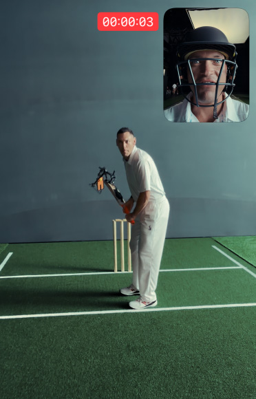 Cricketer Scott Boland stands in front of a wicket on an indoor cricket pitch holding a bat with iPhone 17 Pro attached, ready to strike. In the top right-hand corner, a picture-in-picture window shows a selfie view of cricketer Alex Carey wearing a helmet and talking.