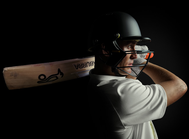 Close-up of cricketer Marnus Labuschagne wearing a helmet and looking ahead with his arm swung to the left, cricket bat held over his shoulder. Shot in low-light against a black background for dramatic effect. 