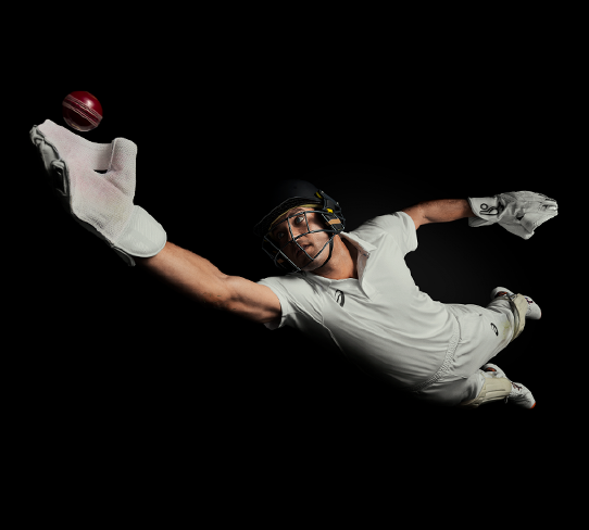 Cricketer Alex Carey reaches upwards with one gloved hand outstretched to catch a red cricket ball, shot from above and at an angle against a black background.