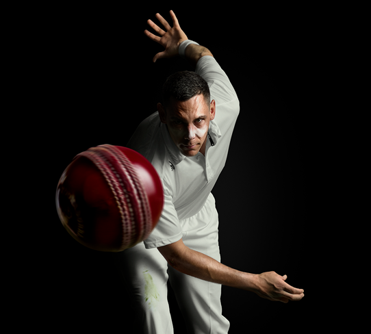 Cricketer Scott Boland bowls a ball directly at the camera. The ball appears at close range suspended in mid-air. Scott leans forwards with one arm outstretched behind him, indicating an action shot.