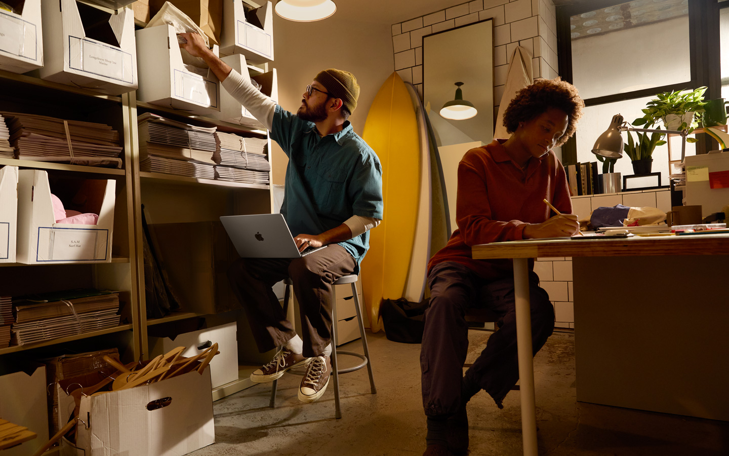 Una persona sentada en un escritorio usa un lápiz para escribir en un cuaderno, otra persona sentada junto a un estante tiene una MacBook en el regazo y estira el brazo derecho para alcanzar un archivo.