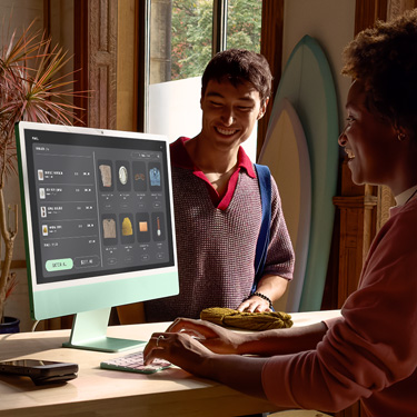 Sörf tahtaları ve başka ürünler satan bir mağazada tezgahın arkasında oturan bir çalışan, iMac kullanarak tezgahın diğer tarafındaki müşterinin satın alma işlemini tamamlıyor.