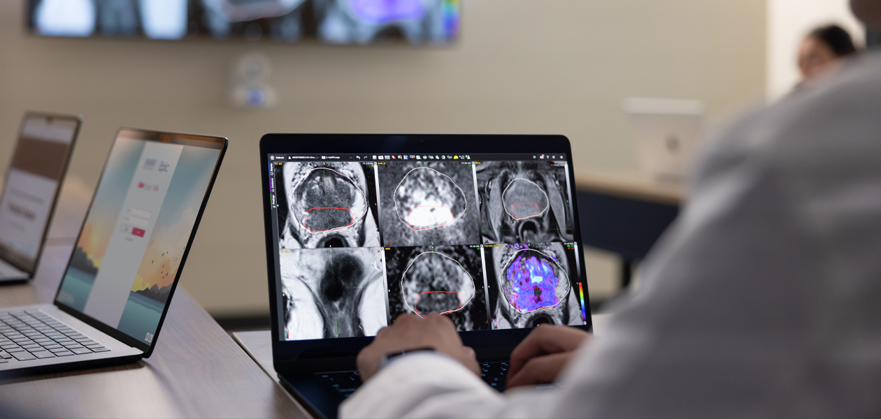 A person reviewing medical scans on a MacBook. Two additional MacBook devices sit open in the background.