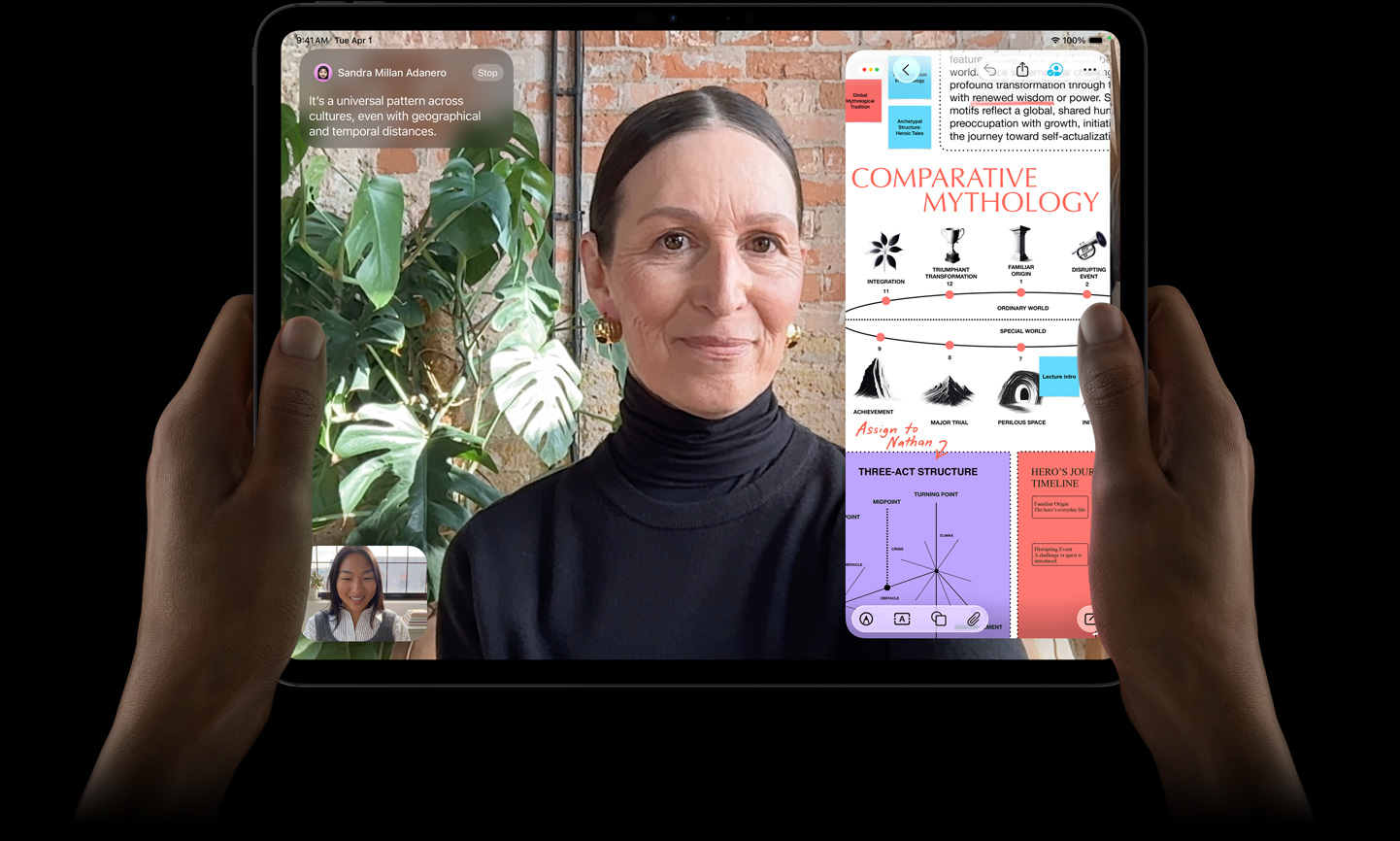 iPad Pro, pohľad spredu, kozmická čierna farba, ľavá a pravá ruka drží zariadenie v orientácii na šírku, zaoblené rohy, čierny rámček displeja, na displeji sa zobrazuje konverzácia cez FaceTime prekladaná v reálnom čase a vzdelávacia apka, v ktorej sa zobrazuje prezentácia „Komparatívna mytológia“ s textom, diagramami a ručne písanými poznámkami