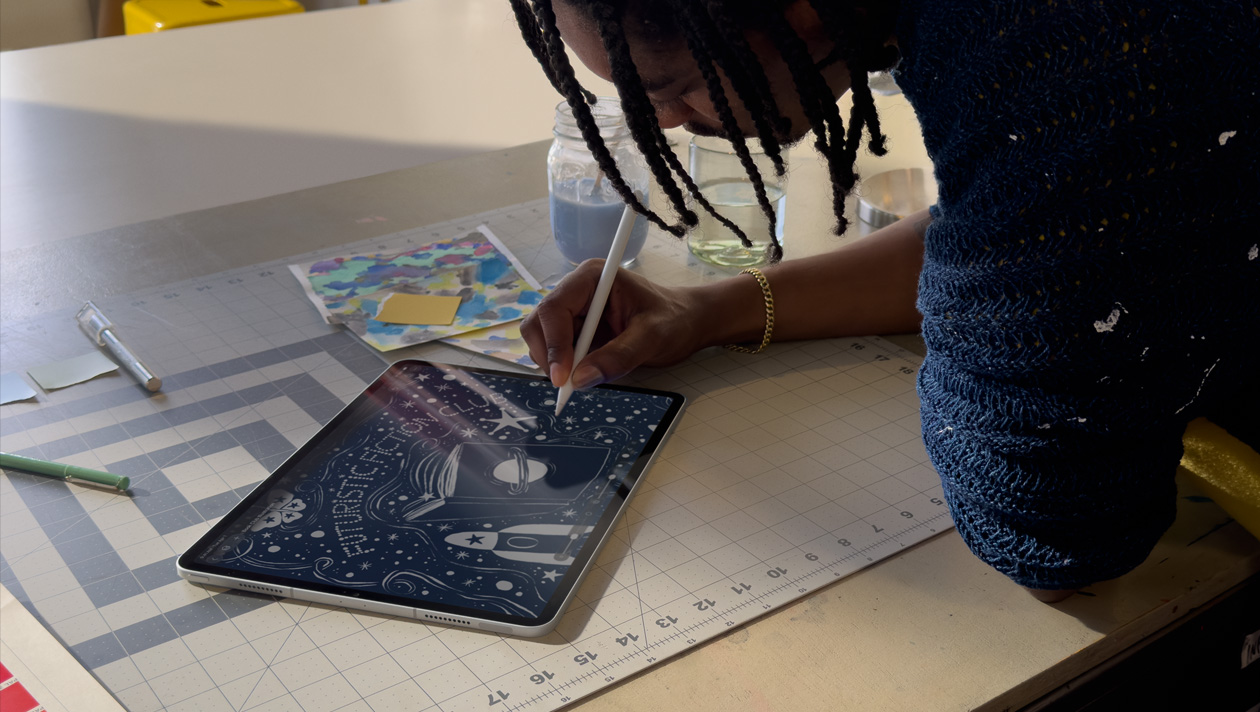 Person with braids wearing a blue sweater drawing on iPad Pro with Apple Pencil, screen shows illustration of a night sky with a planet on a book cover, space ship, butterfly and futuristic fiction club written in the centre